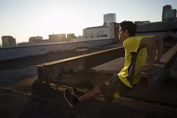 A man in exercise clothes on a rooftop overlooking the city, doing bench shoulder push ups.
