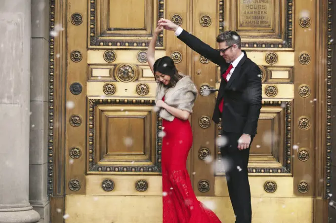 A woman in a long red evening dress with fishtail skirt and a fur stole, and a man in a suit dancing on the steps of a building.