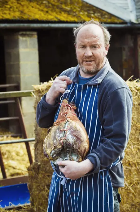 Butcher wearing a striped blue apron, standing outdoors, holding a large cured ham.