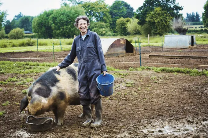 A woman stood by a large pig holding a bucket.