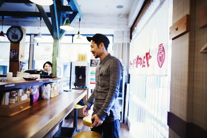 A ramen noodle cafe in a city.  Customer standing up at the counter ordering food.