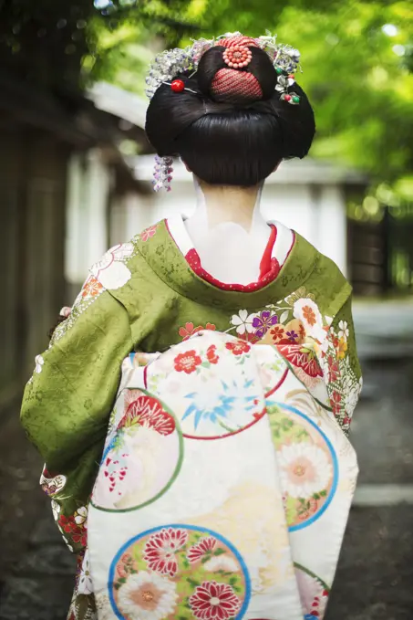 A woman dressed in the traditional geisha style, wearing a kimono and obi, with an elaborate hairstyle and floral hair clips, with white face makeup with bright red lips and dark eyes on a street.