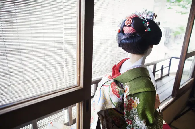 A woman dressed in the traeditional geisha style, wearing a kimono and obi, with an elaborate hairstyle and floral hair clips, with white face makeup.