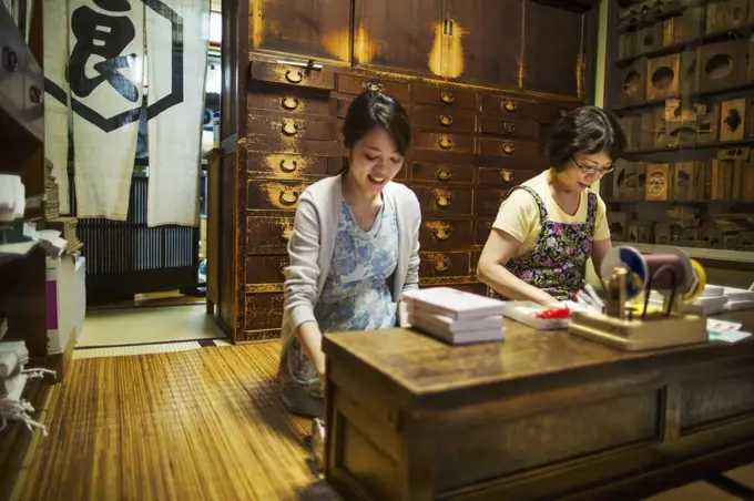A traditional wagashi sweet shop. A woman working at a desk using a laptop and phone. A woman packing merchandise.