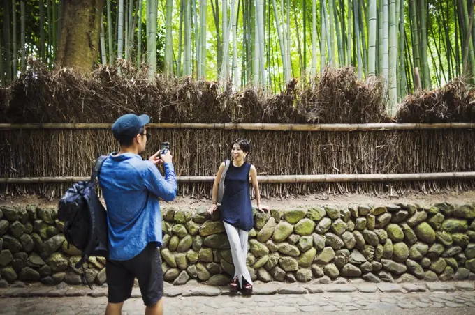 A man taking a photograph of a woman by a fence around woodland.