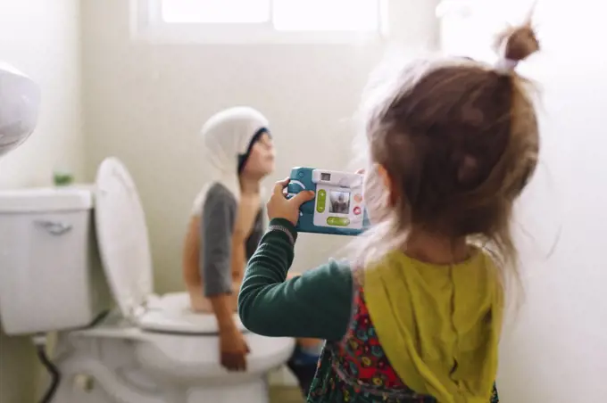 Young girl standing in bathroom, holding camera, taking picture of boy sitting on toilet with his shirt over his head.