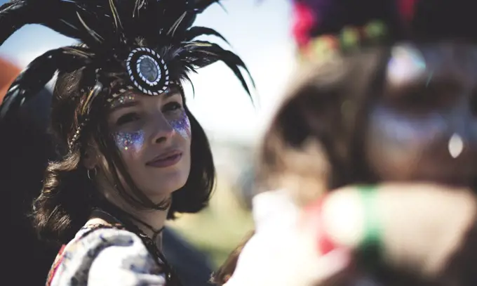 Young woman at a summer music festival face painted, wearing feather headdress, looking at camera.
