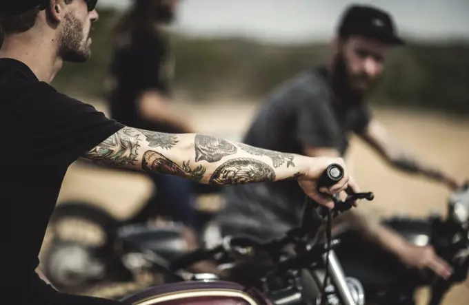 Side view of man with tattoos on his arm sitting on cafe racer motorcycle on a dusty dirt road.