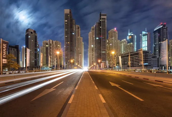 Cityscape with illuminated skyscrapers in Dubai, United Arab Emirates at dusk, highway in foreground.