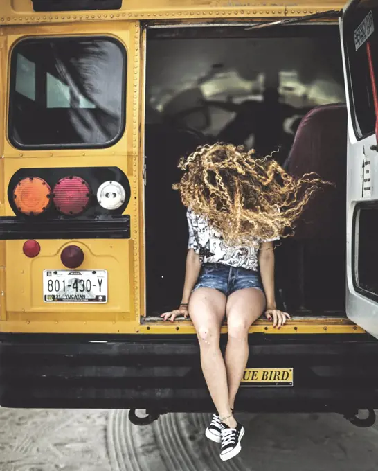 A young woman with long curly hair over her face sitting on the tailgate of a school bus.