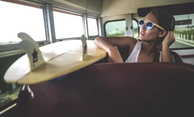 Young woman sitting on a bus with a surfboard.