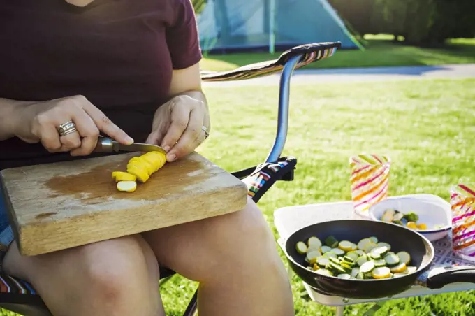 A person sitting beside a camp stove preparing yellow zucchini, courgettes for the frying pan.