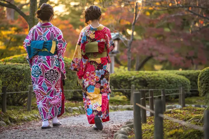 Rear view of two women wearing colourful traditional Japanese kimonos walking along a path in a park.