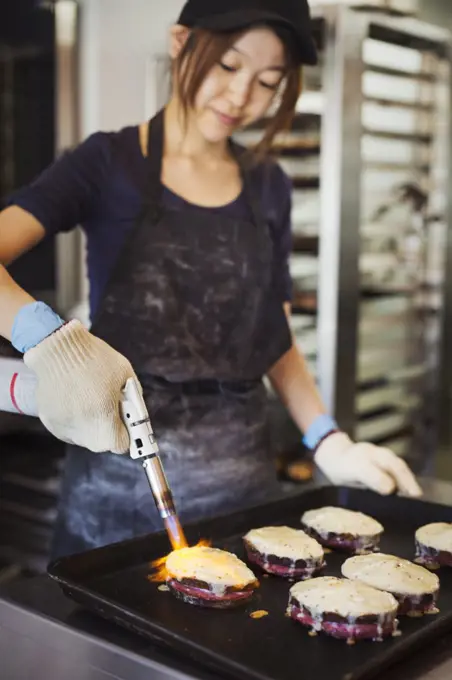 Woman working in a bakery, wearing oven gloves, using blowtorch, melting cheese on sandwiches.