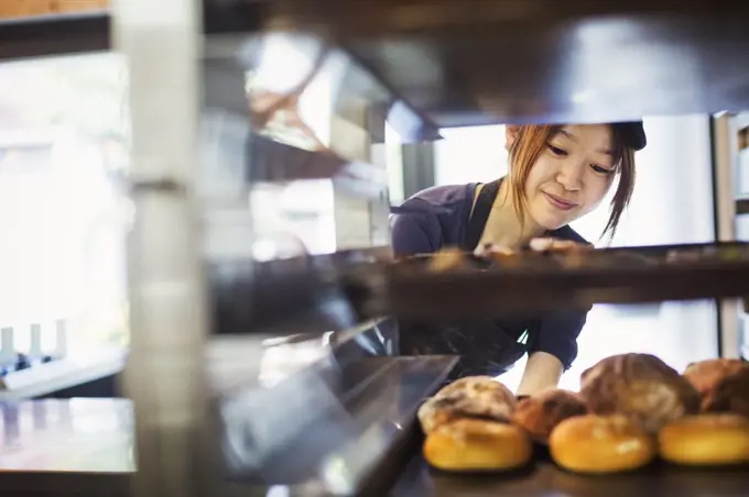 Woman working in a bakery, placing large trays with freshly baked rolls on a trolley.
