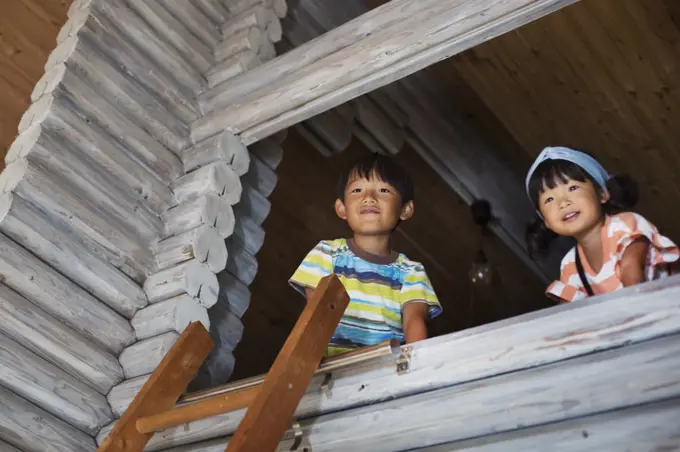 Low angle view of boy and young girl with black hair looking out of window, ladder leaning against wooden wall.