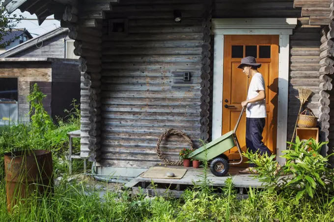 Man wearing hat standing outside wooden garden shed, pushing green wheelbarrow.