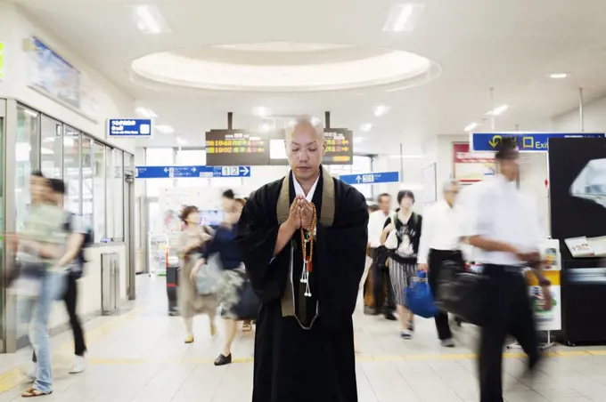 Buddhist monk with shaved head wearing black robe standing inside a train station, holding mala, people walking past.
