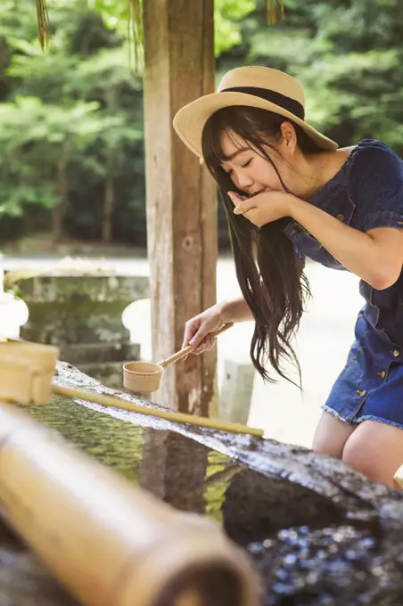 Young woman wearing blue dress and hat using bamboo water hand washing basins at Shinto Sakurai Shrine, Fukuoka, Japan.