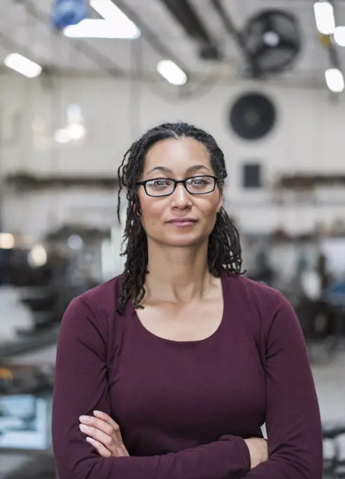 Woman with brown hair wearing glasses standing in metal workshop, smiling at camera.