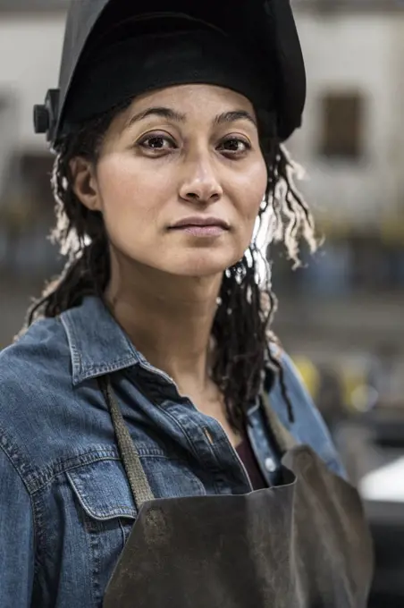 Portrait of woman wearing apron and welding mask standing in metal workshop, smiling at camera.