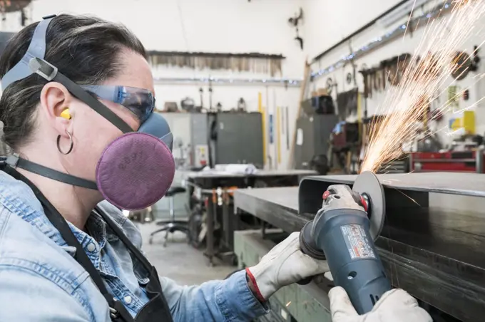 Woman wearing safety glasses and dust mask standing in metal workshop, using power grinder, sparks flying.