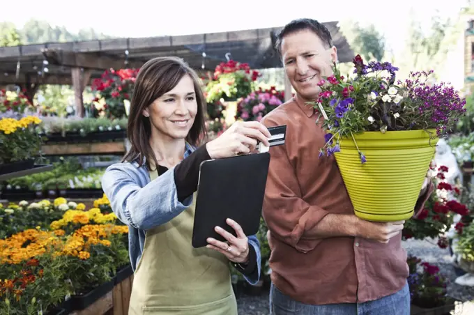 Caucasian man buying plants with a credit card at a nursery owned by a Caucasian woman using a credit card reader on a notebook computer