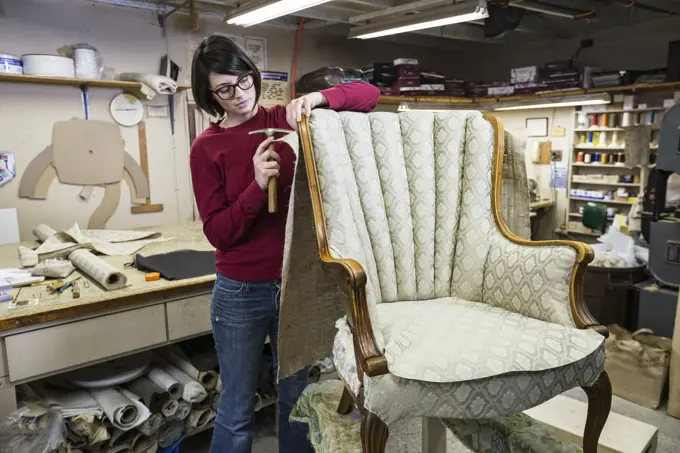 A young female upholsterer using a tack hammer on a chair in an upholstery shop.