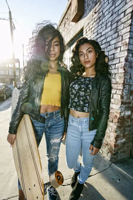 Two young smiling women with long curly black hair standing on pavement, holding skateboard, looking at camera.