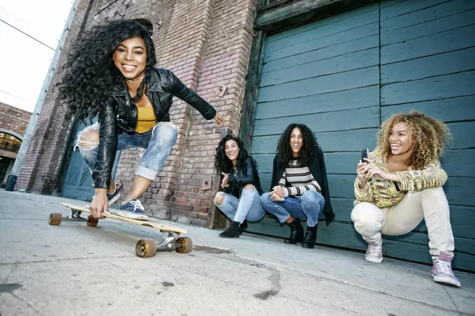 Three young women with curly hair squatting in front of shutter, watching smiling young woman riding a skateboard.