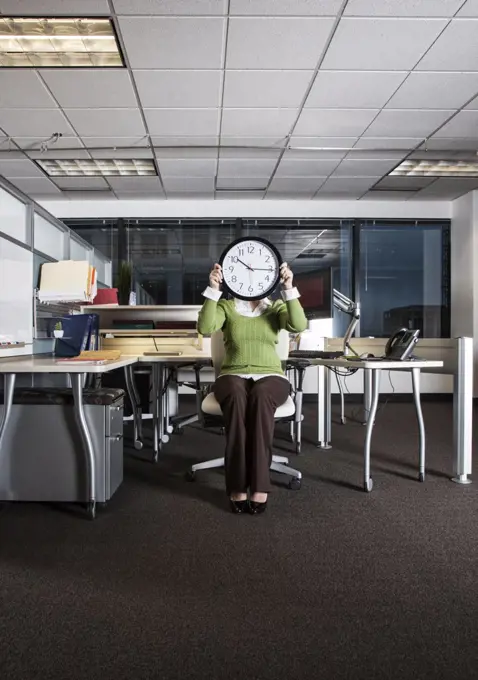Woman holding a clock face while sitting in her cubicle office space.