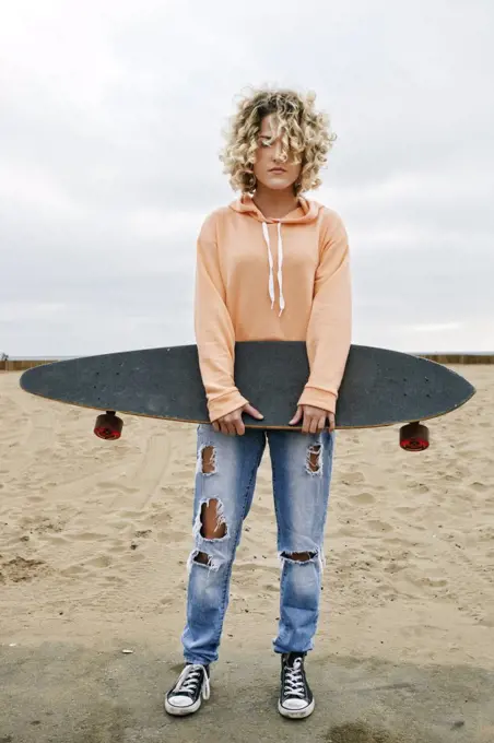 Young woman with curly blond hair wearing pink hoodie and ripped jeans standing on sandy beach, holding skateboard, looking at camera.