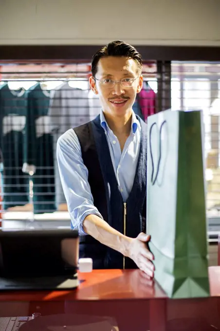 Japanese salesman with moustache wearing glasses standing at counter in clothing store, holding green shopping bag, smiling at camera.
