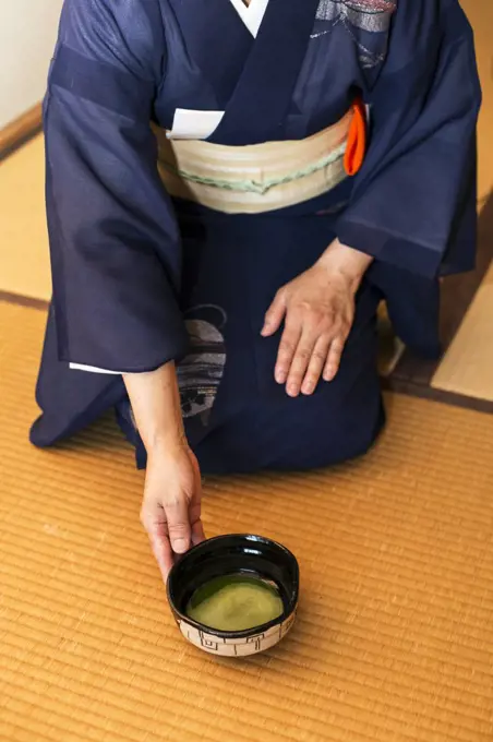 High angle view of Japanese woman wearing traditional bright blue kimono with cream coloured obi kneeling on floor, holding bowl with Matcha tea.
