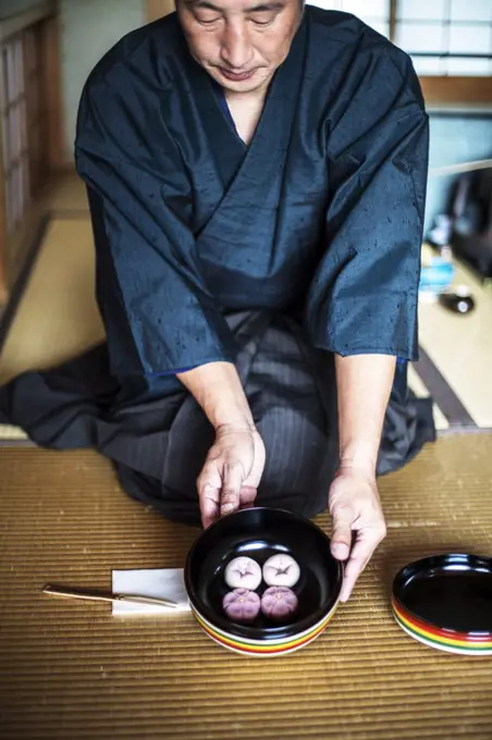Japanese man wearing traditional kimono knelling on floor, holding a bowl with Wagashi, sweets traditionally served during a Japanese Tea Ceremony.