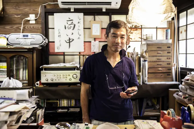 Japanese man standing in a Washi producing workshop holding a smart phone, smiling at camera.