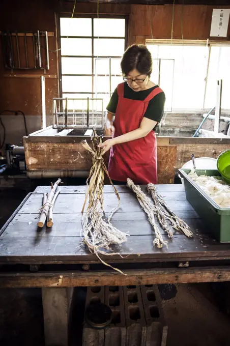 Woman standing at a table in a traditional Washi workshop, stripping bark and fibre off twigs and branches of plant material 
