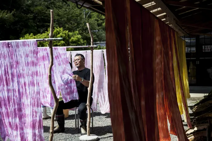 Japanese man sitting outside a textile plant dye workshop, hanging up freshly dyed pink fabric.