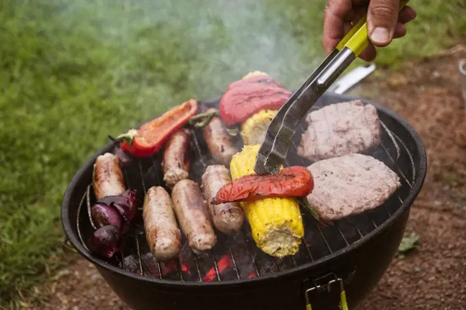 A person using tongs on food on a barbeque, sausages and burgers, sweetcorn and red peppers cooking over glowing hot coals.