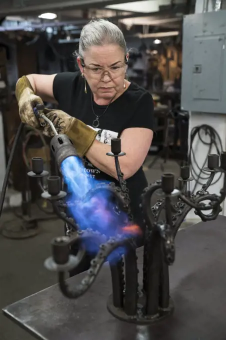 Caucasian female blacksmith using a torch to heat a metal candelabra in her studio.