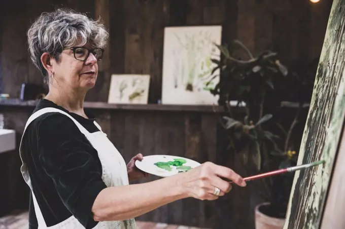 Senior woman wearing glasses, black top and white apron standing in studio, working on painting of trees in forest.