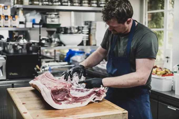 Male butcher wearing apron and black rubber gloves cutting pork ribs on butcher's block.