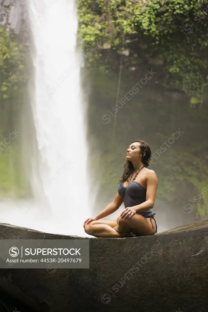 Woman meditating on rock near waterfall in jungle