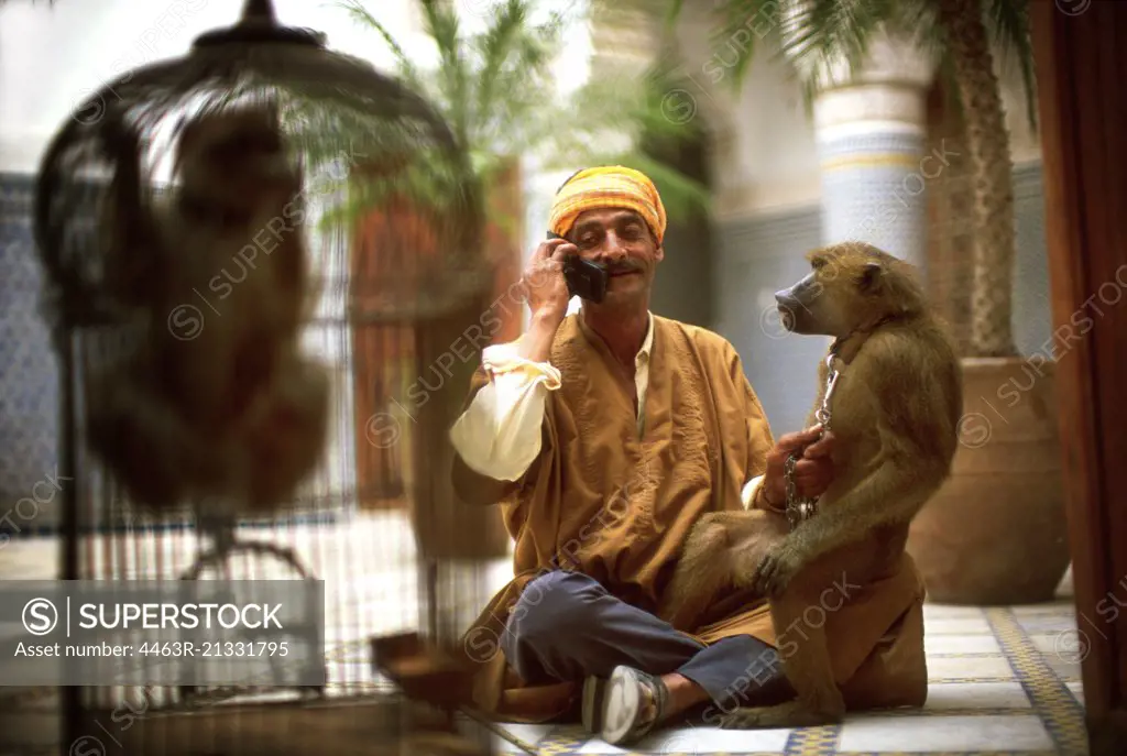 Mid-adult man sitting in a courtyard with a monkey on his knee as he talks on a cell phone.