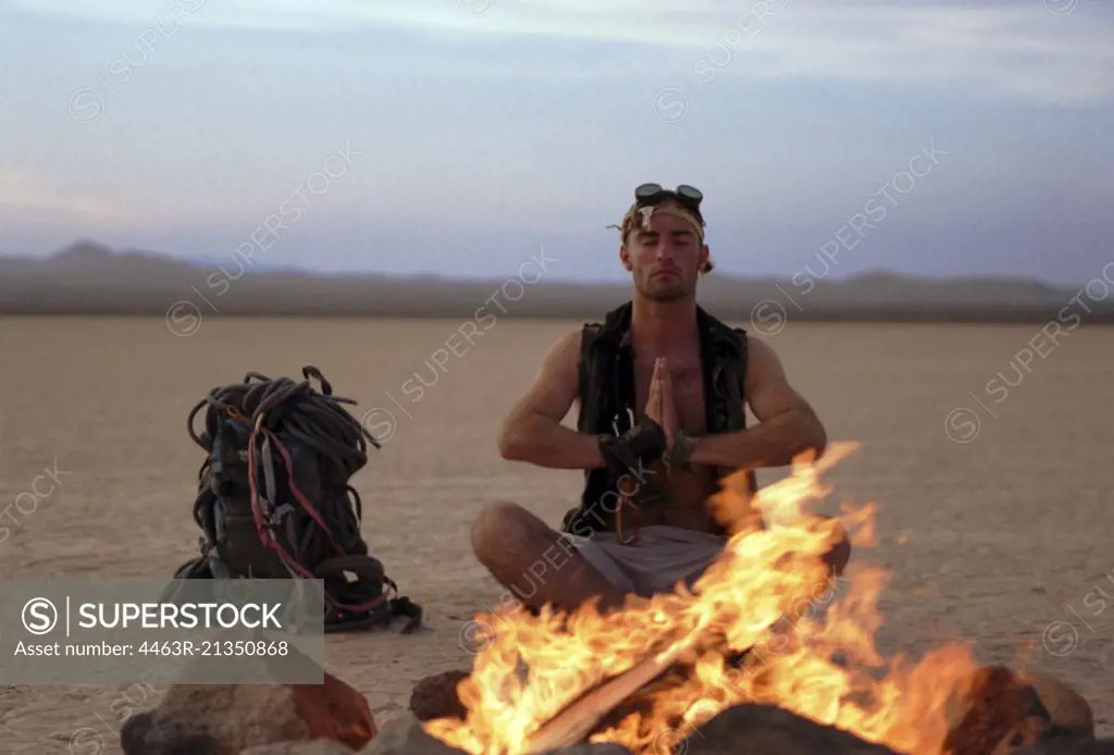 Young man sitting meditating with his eyes closed in front of a campfire in the desert.