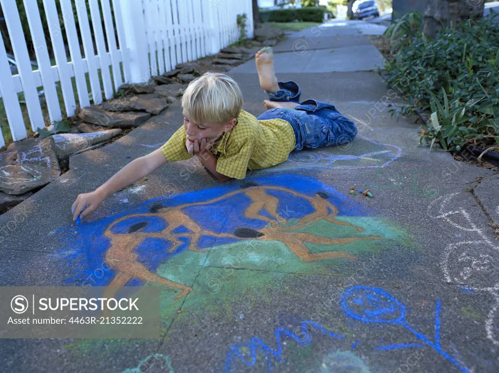 Young boy lying on his stomach and drawing on a suburban sidewalk outside his house.