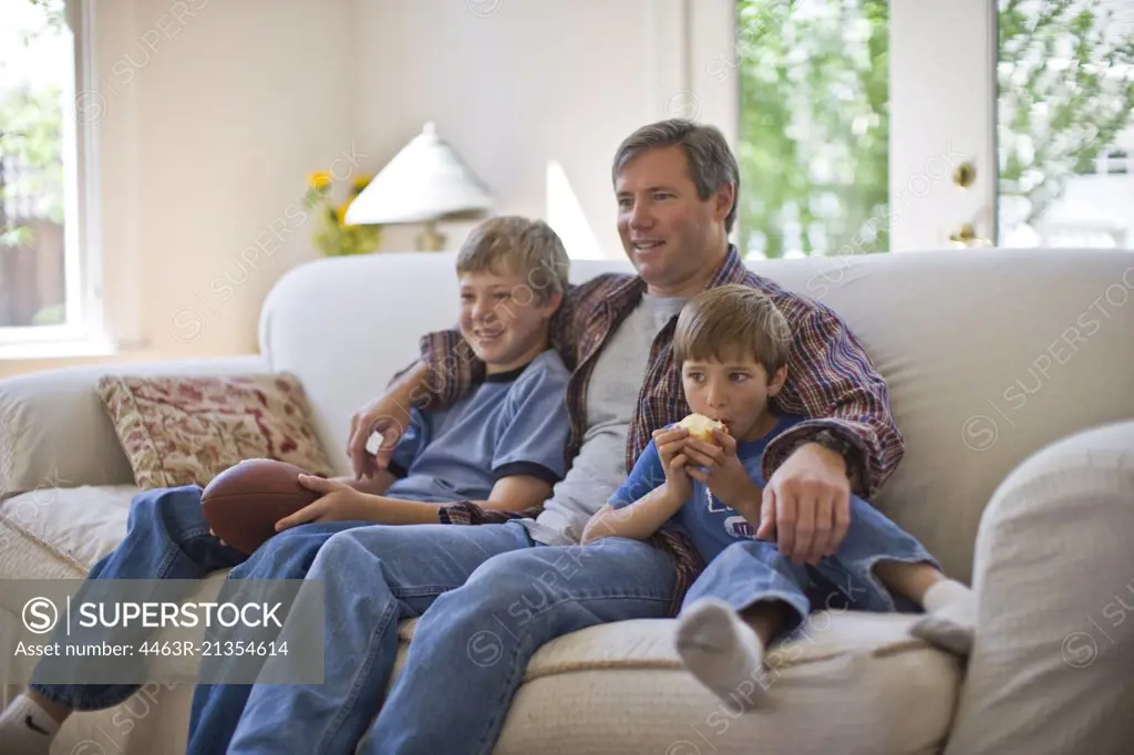 Mid-adult man sitting with an arm around his two young sons on a sofa in their living room.