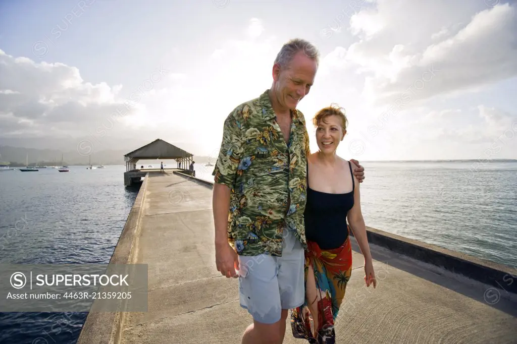 Happy heterosexual couple smile as they walk together along a jetty with their arms around each other.