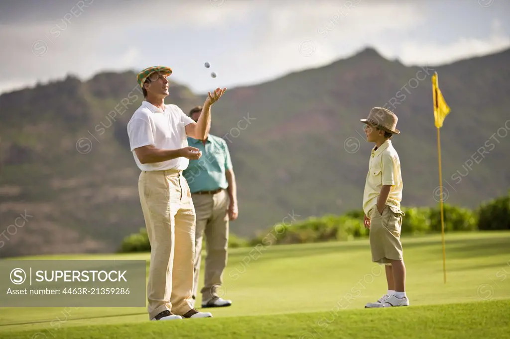 Young boy watches his golfer father juggle golf balls as they stand on a green.