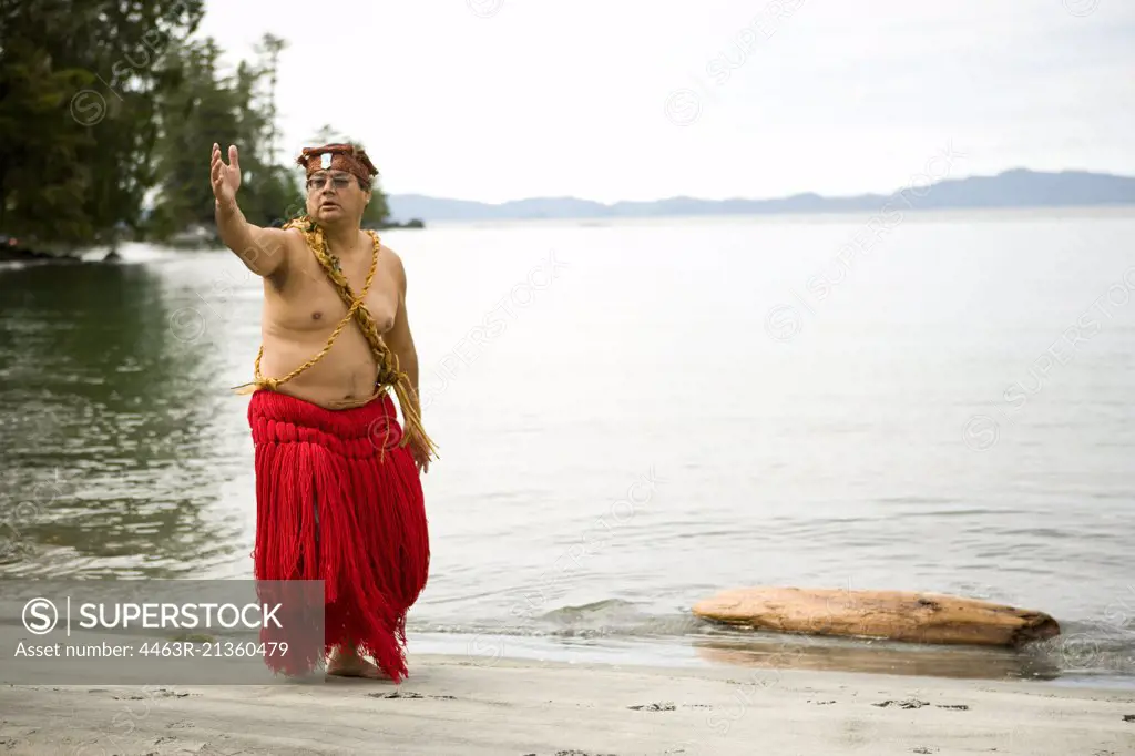 Mid-adult man wearing traditional dress performing a cultural dance beside a lake.